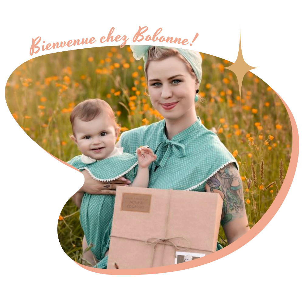 femme habillée en pinup tient un bébé et une box en kraft dans les bras. Elles sont toutes les deux très souriantes.
